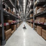 An Asian woman sits in a large warehouse aisle filled with inventory racks and shelves.